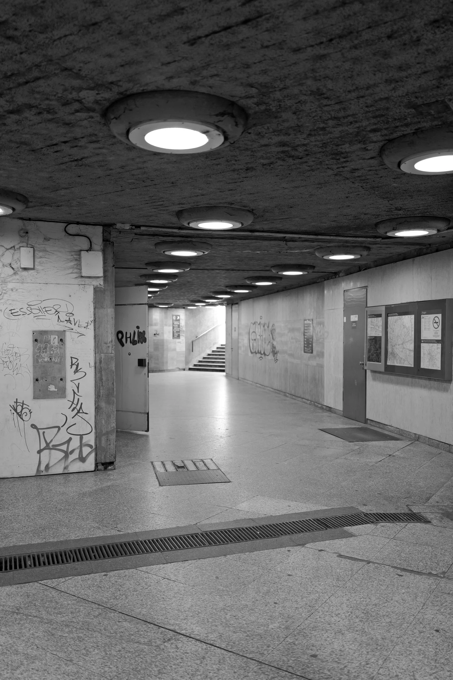 Passage souterrain avec plafonds éclairés par des sièges encastrés, murs en béton avec graffiti, panneaux d'information et escaliers à l'arrière.