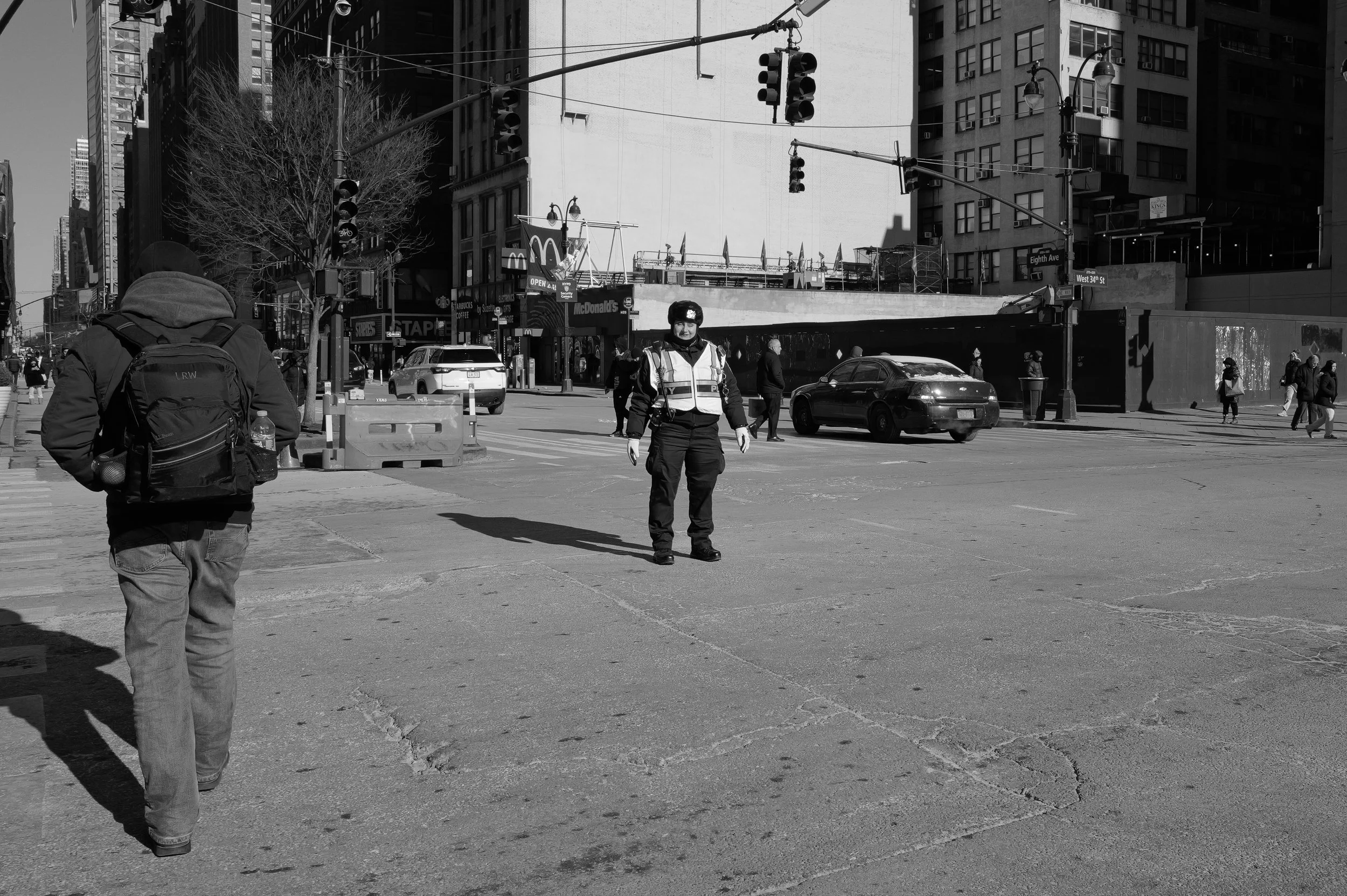 Un agent de police en uniforme et en gilet de sécurité se tient au centre d'une intersection urbaine, entouré de quelques piétons et voitures, avec des gratte-ciel en arrière-plan.