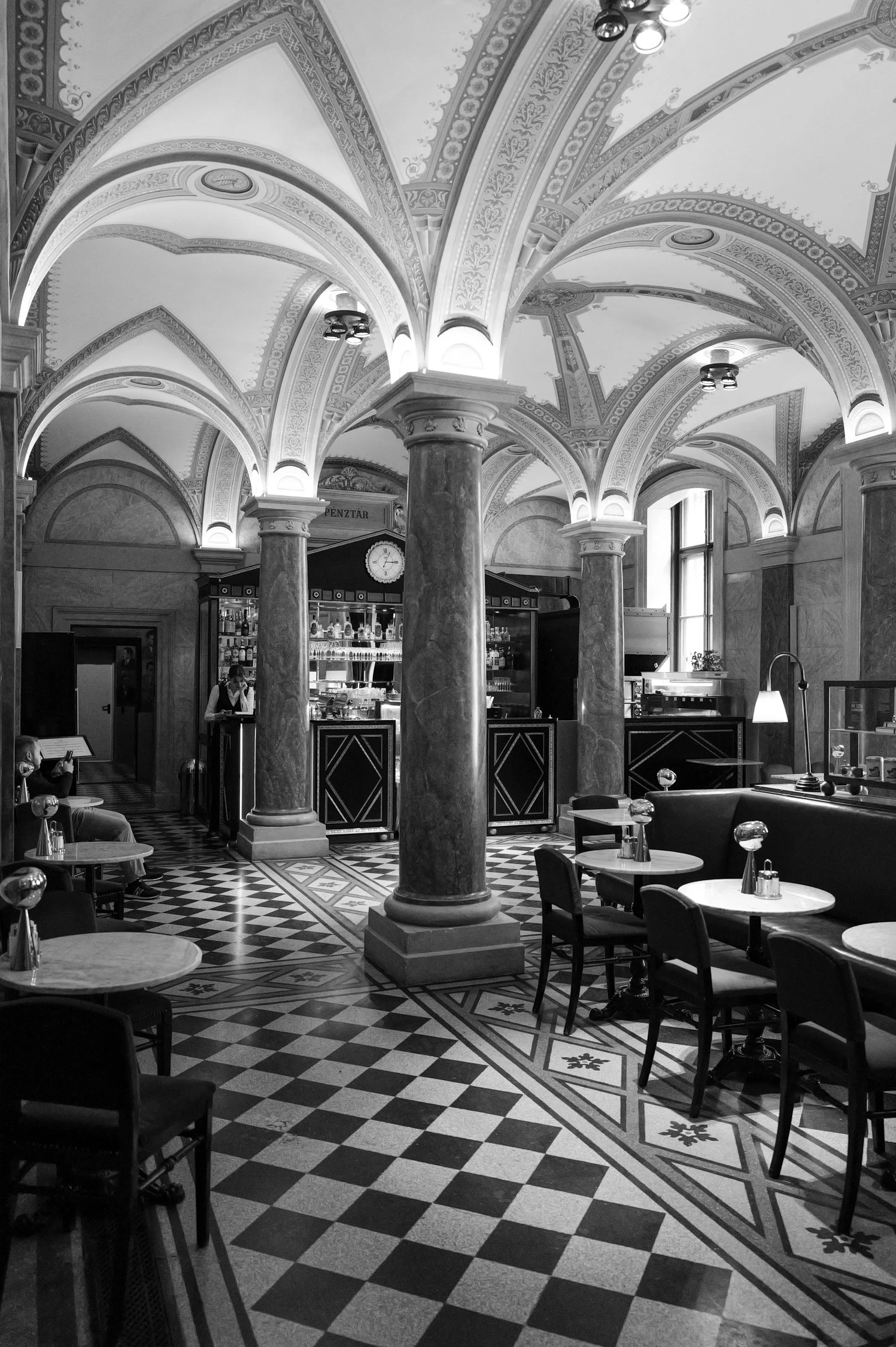 Une salle élégante avec des plafonds voûtés ornés de décorations détaillées, plusieurs colonnes en marbre, un comptoir de bar, des tables rondes avec des chaises, éclairée par des fenêtres et plafonniers.