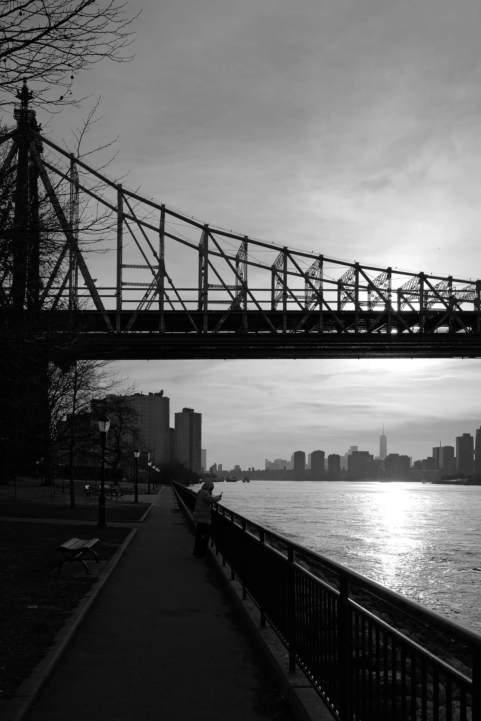 Une femme marche le long d'une promenade au bord de la rivière avec une vue sur la ville et un pont en acier en arrière-plan, en noir et blanc.