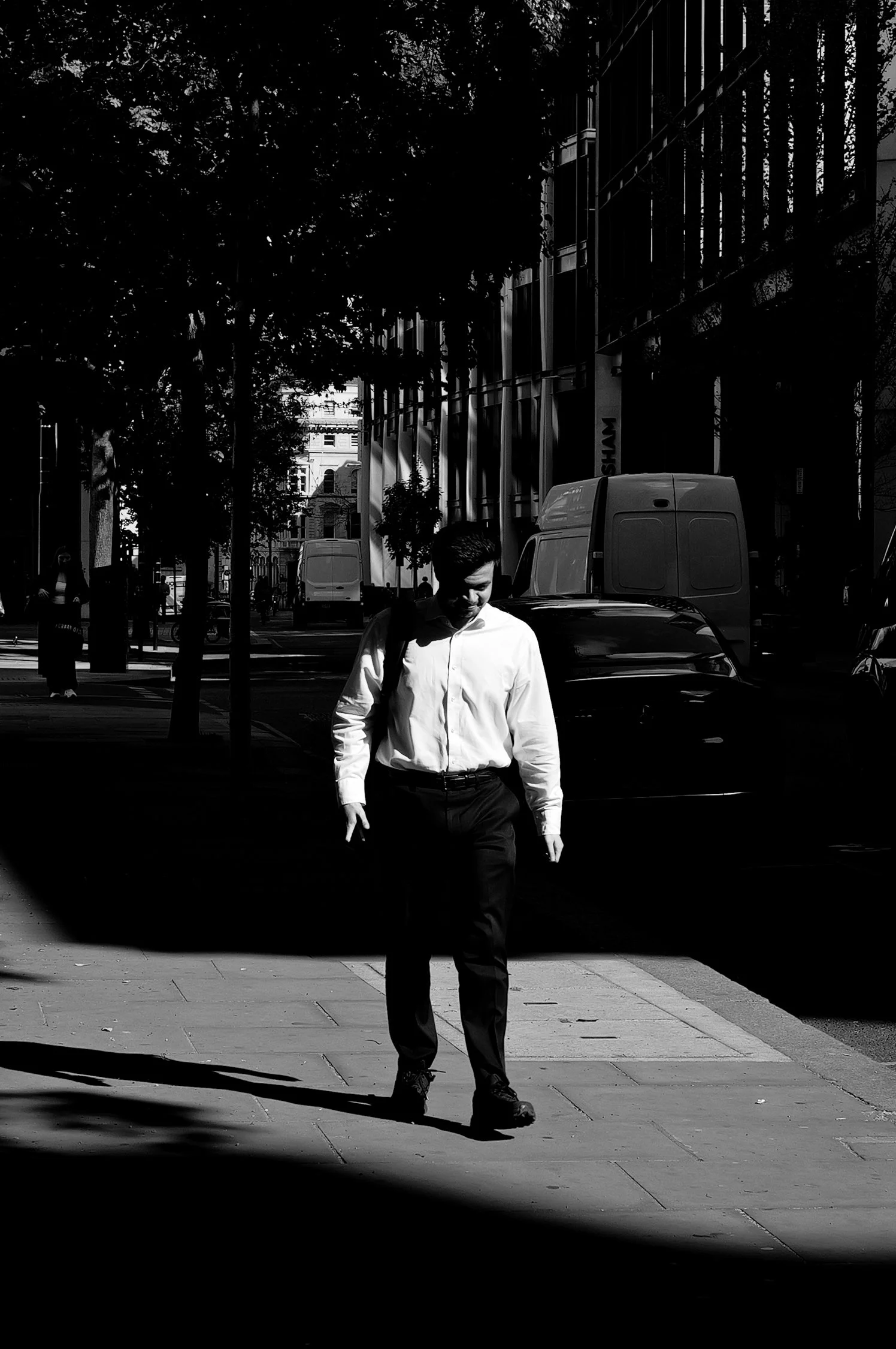 Un homme en chemise blanche marche sur un trottoir d'une rue urbaine, avec des bâtiments modernes et des véhicules en arrière-plan, dans une ambiance ensoleillée.