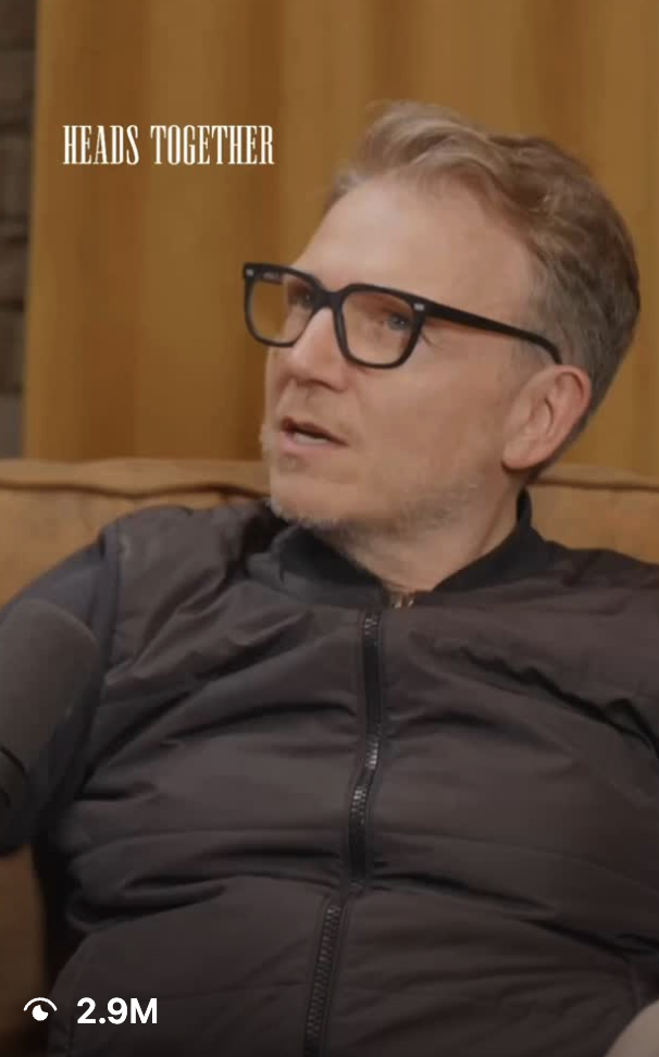 A man with light brown hair, glasses, and wearing a black jacket, sits on a brown leather sofa in a room with yellowish wood paneling. There is text that says "HEADS TOGETHER" in the upper left corner.