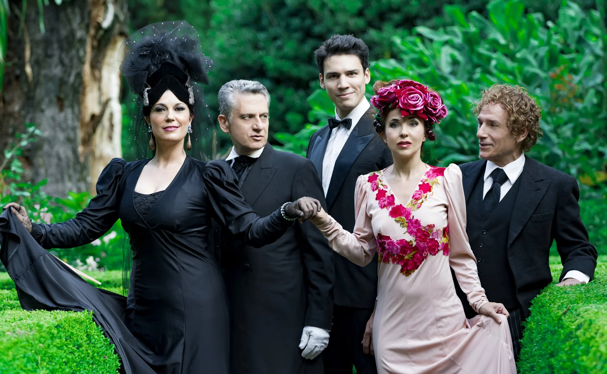 Group of six people dressed in vintage formal clothing, standing outdoors in a lush garden with green trees and bushes in the background.