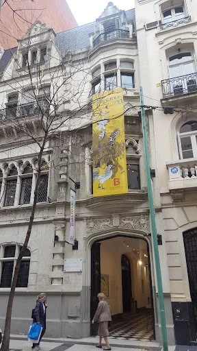Historic multi-story building with ornate architecture, a yellow banner hanging from the facade, and pedestrians walking on the street.