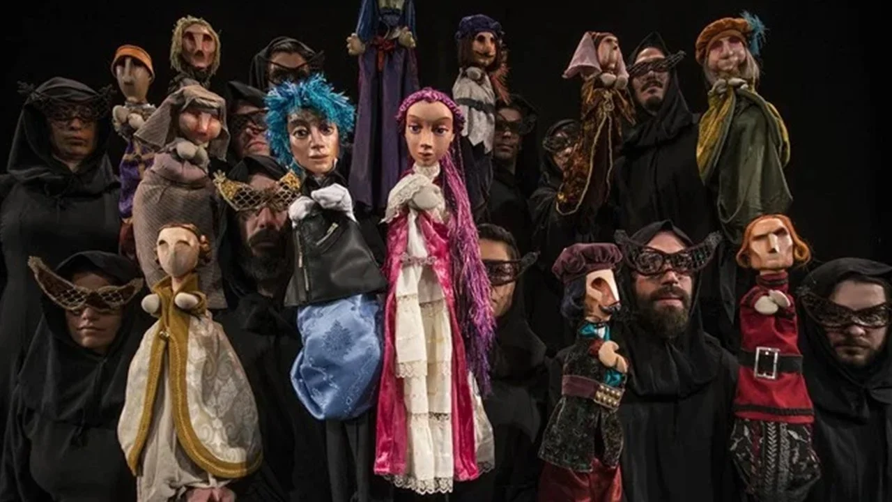 Group of people wearing black hooded cloaks and puppet masks, some with colorful hairstyles and elaborate costumes, holding puppet characters.