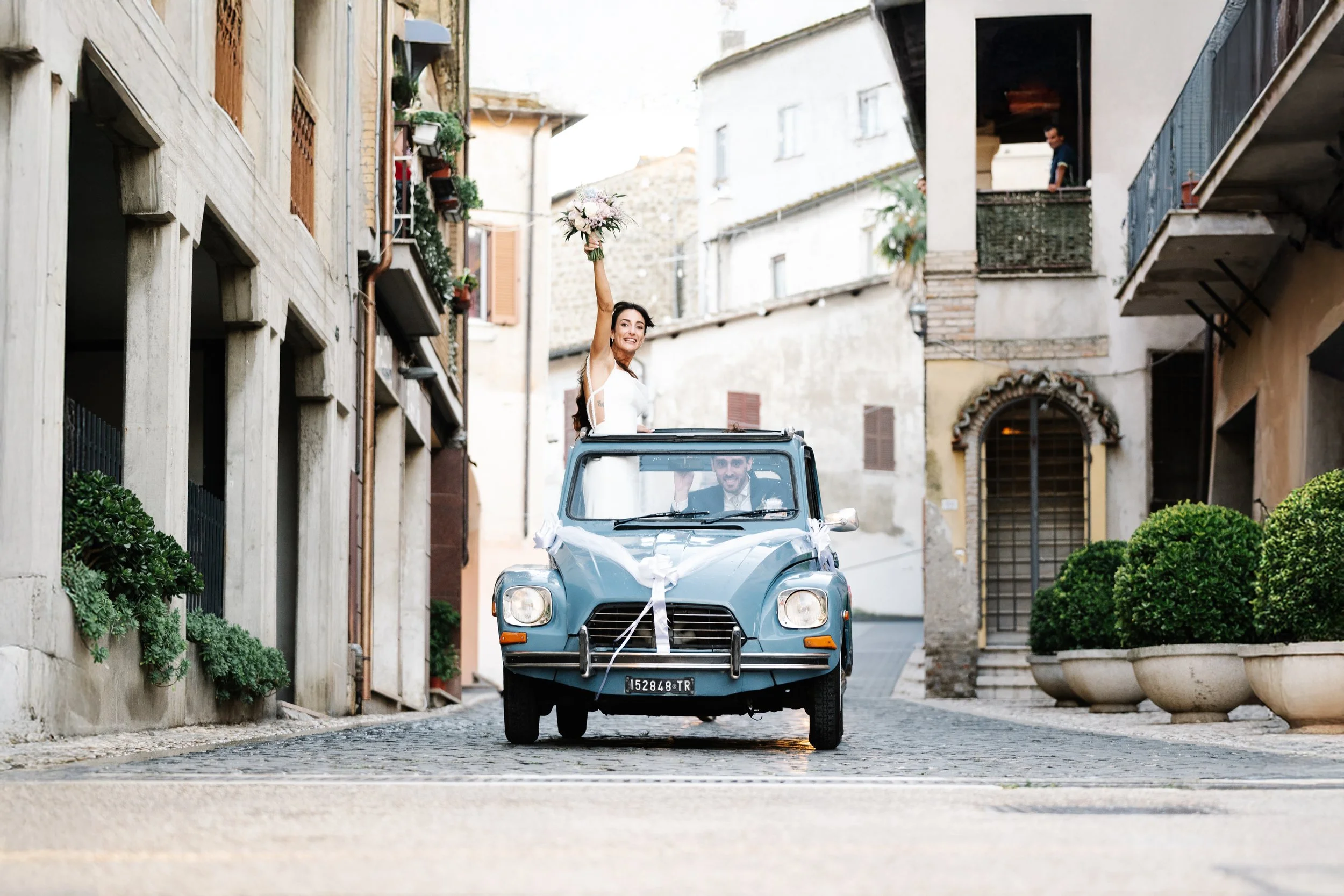 A bride and groom riding in a vintage blue car decorated with white ribbons on a European narrow street, with the bride holding a bouquet and smiling, while the groom drives.