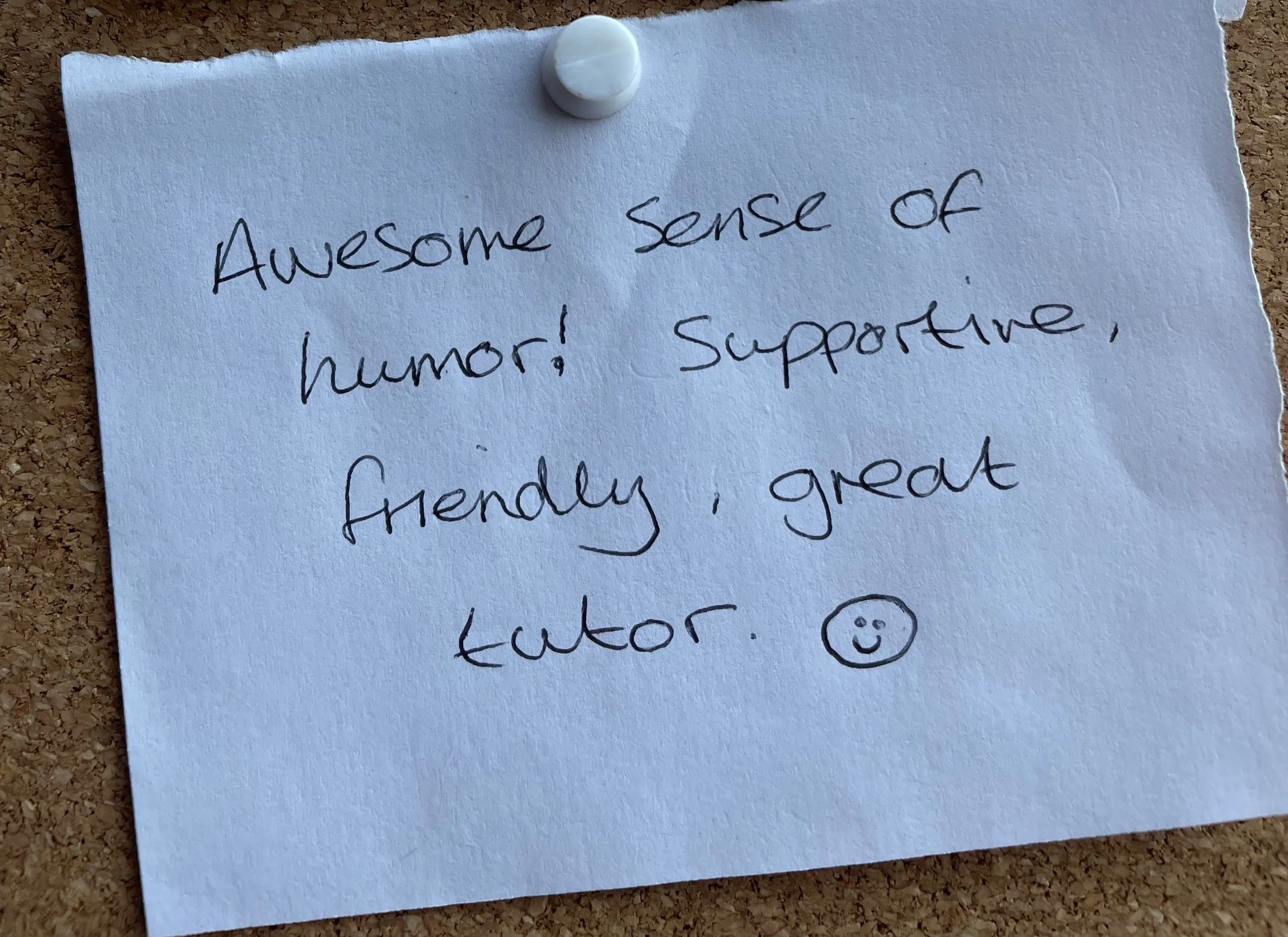 A handwritten note pinned to a corkboard, expressing that it has an awesome sense of humor, supportive, friendly, and great tutor, with a smiley face drawing.