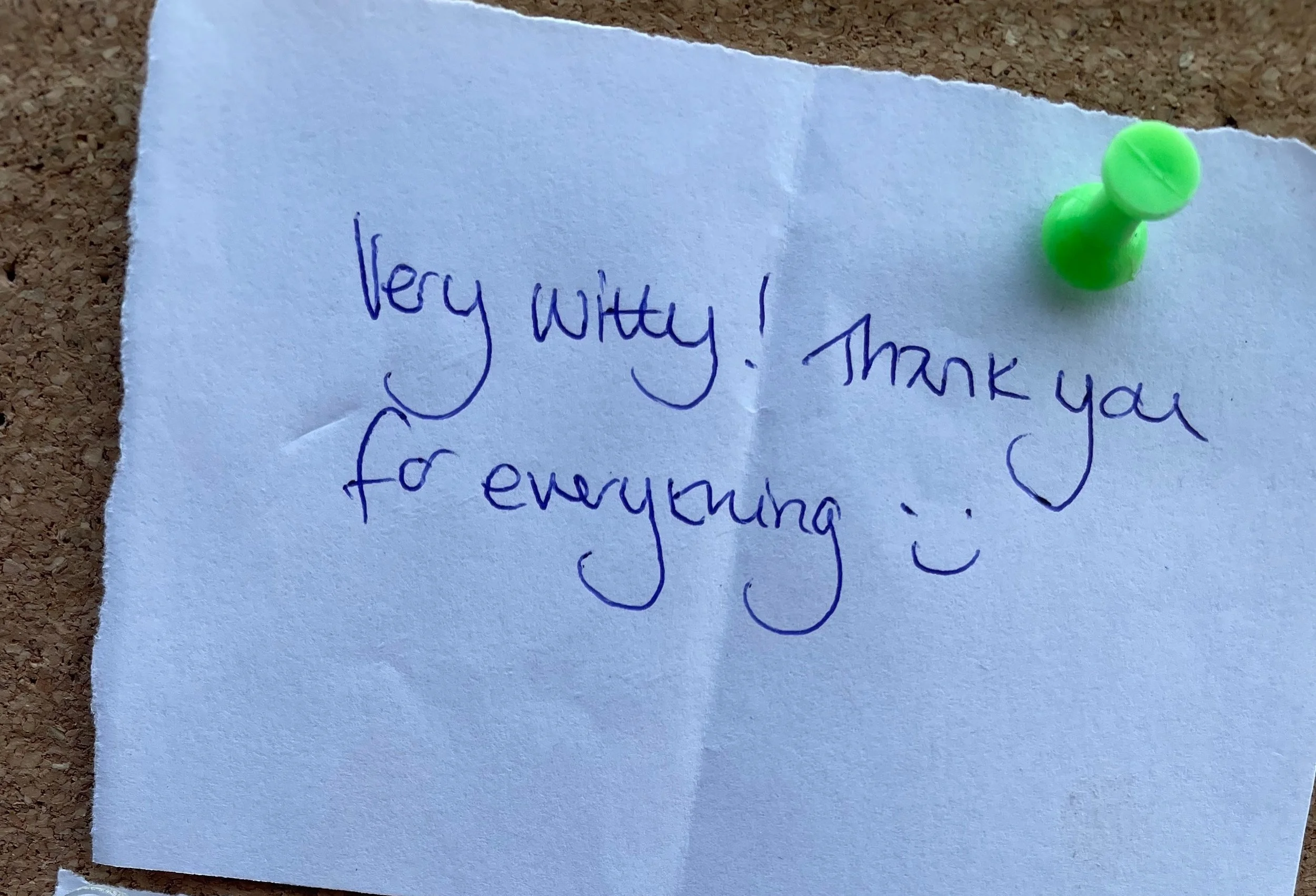 A handwritten note on a piece of paper pinned to a corkboard with two green pushpins. The note reads, "Very witty! Thank you for everything :)" in blue ink.