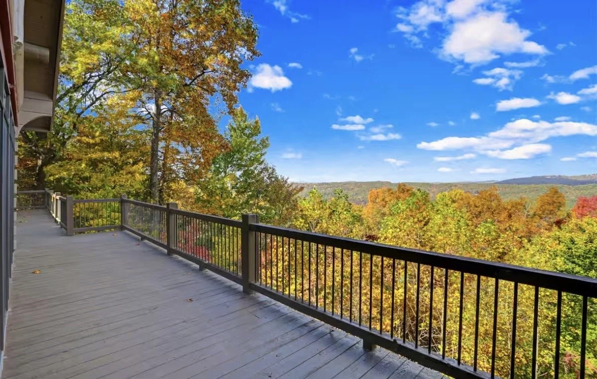 A balcony with wooden flooring and black metal railing, overlooking a landscape of trees with autumn foliage and a blue sky with scattered clouds.