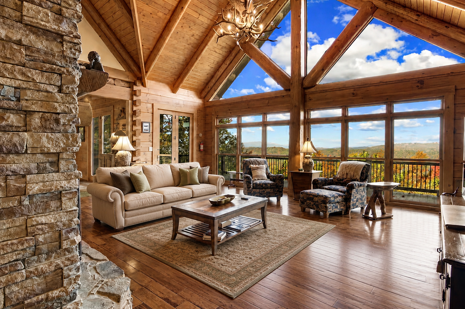 Living room with wooden walls and ceiling, large windows with a mountain view, beige sofa, patterned armchairs, a wooden coffee table, lamps, and a stone fireplace.