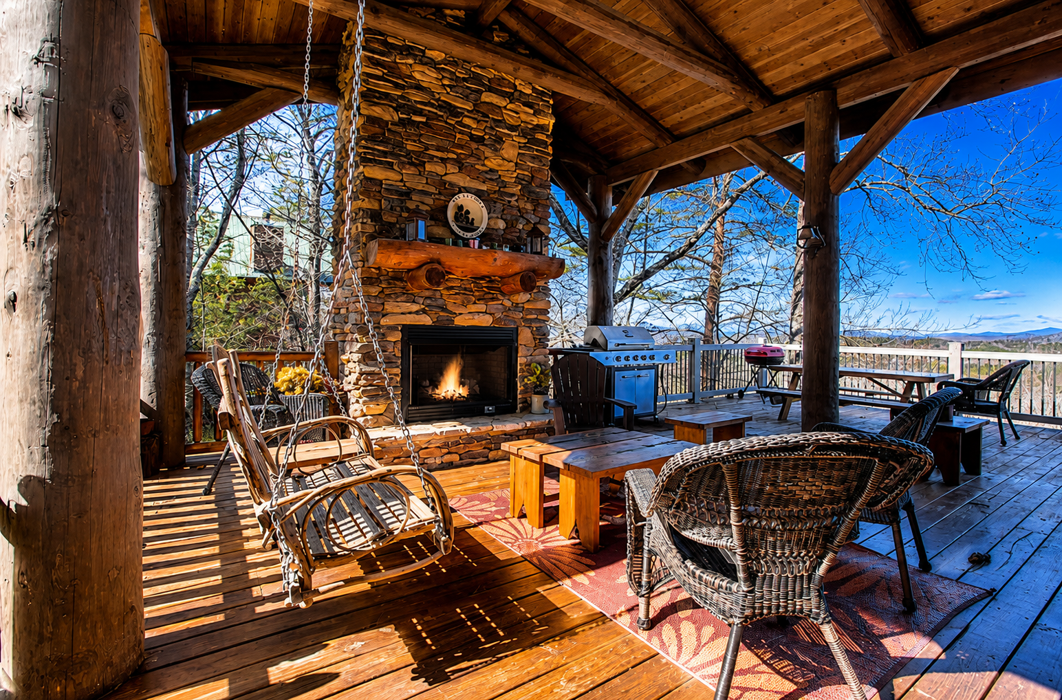 Outdoor deck with a stone fireplace, fireplace flames, wooden ceiling, various outdoor furniture including a swing, chairs, benches, and a barbecue grill, overlooking a scenic view with trees and mountains.