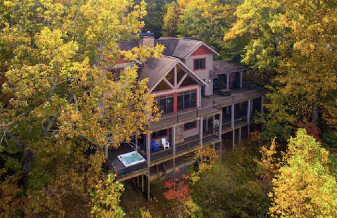 A large house surrounded by colorful autumn trees with yellow, orange, and red leaves. The house has a spacious porch and a hot tub on the deck.