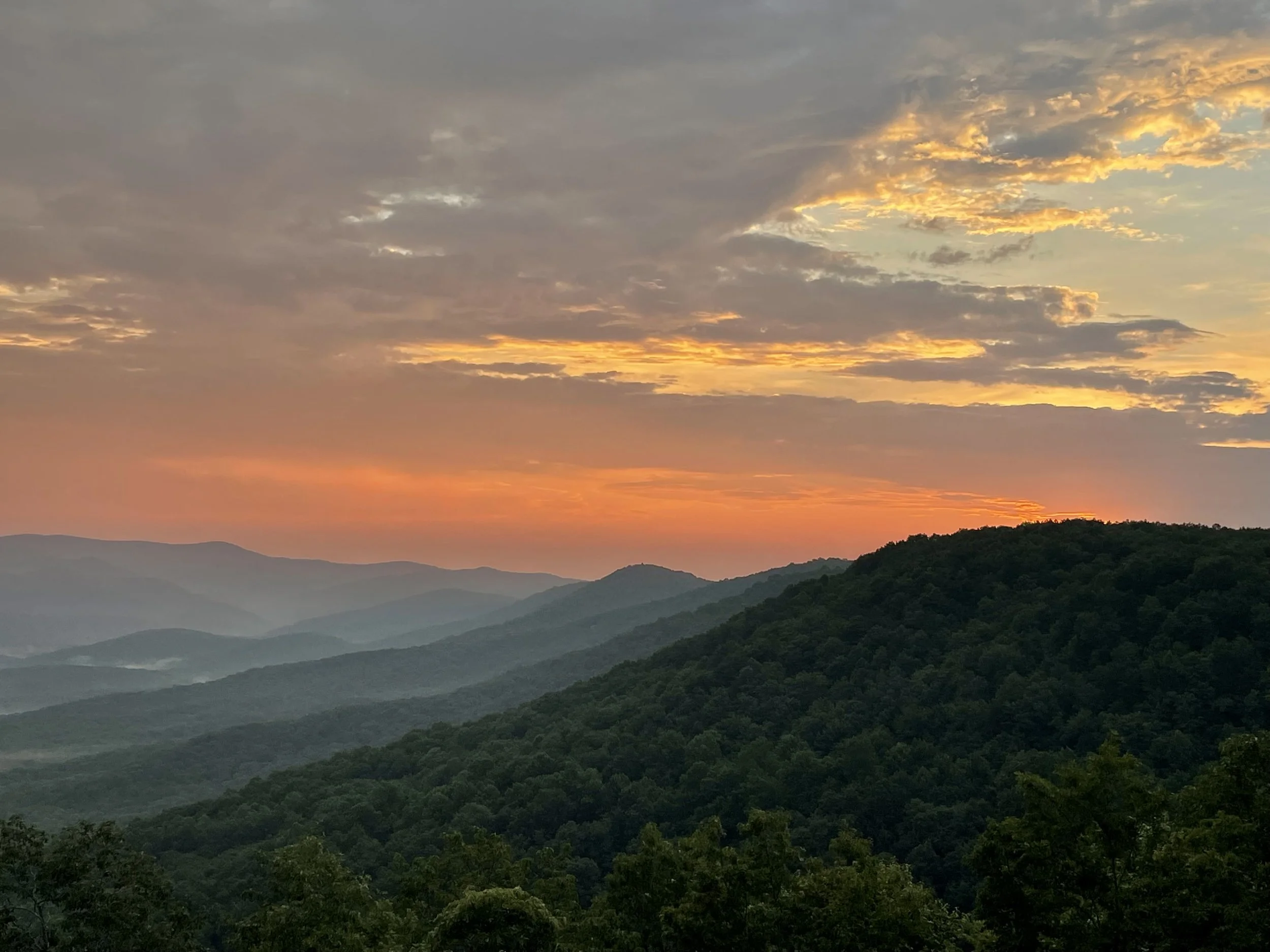 A scenic view of rolling green hills and mountains under a colorful sunset sky with scattered clouds.
