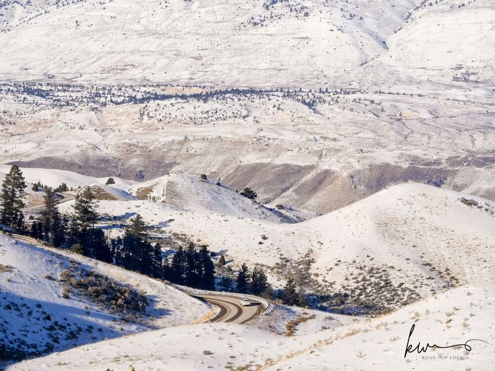white car traveling on clear road surrounded by snowy rolling. hills