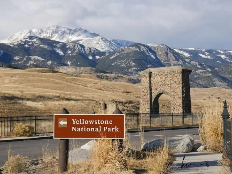 A road sign pointing left to Yellowstone National Park, with a stone arch monument and mountainous landscape in the background.