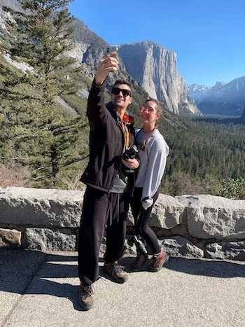 A man and woman taking a selfie in front of a mountain landscape with trees, rocks, and clear blue sky.