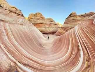 Person standing in the Wave, a colorful sandstone formation with layered, curved patterns in shades of orange, pink, and white, outside under a clear blue sky.
