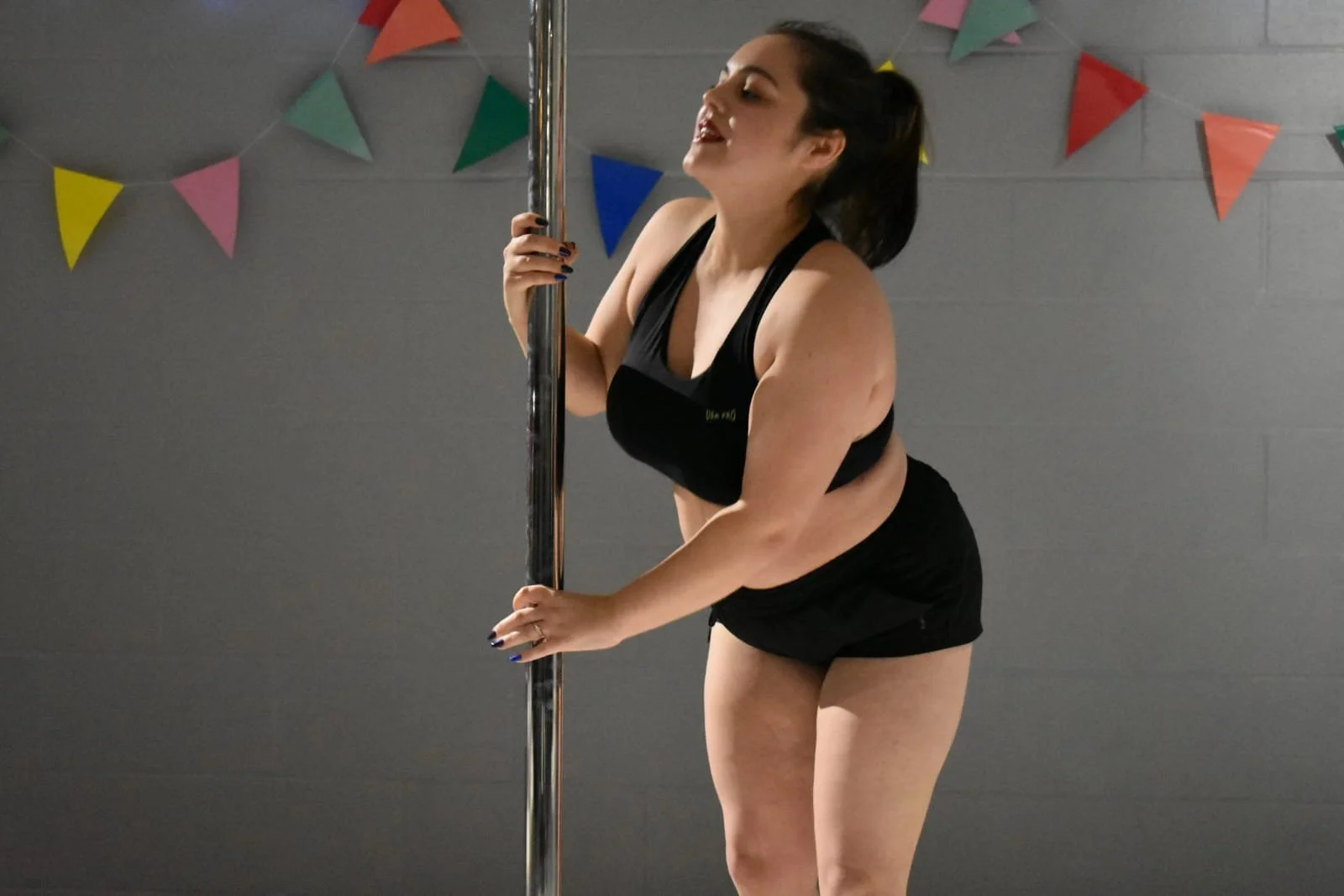 A woman in black sportswear holding a vertical pole in an indoor setting, with colorful triangular banners on the wall behind her.