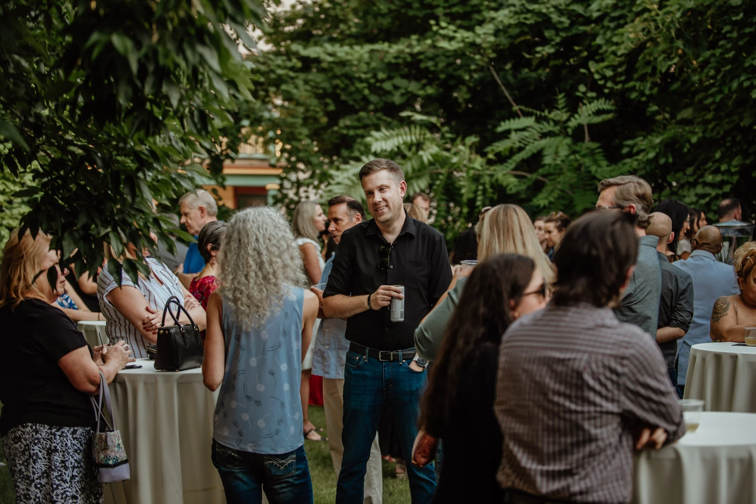 People gathered outdoors at a social event surrounded by greenery, engaging in conversations, some holding drinks, with high-top tables and trees in the background.