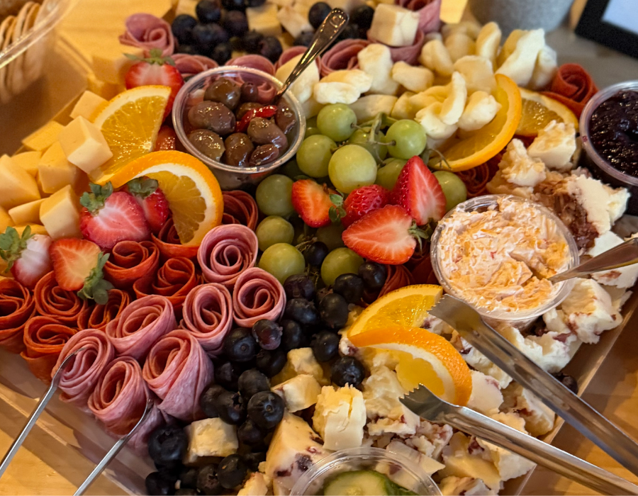 Assorted fruit platter with strawberries, green and purple grapes, orange slices, cheese, rolled meats, and small bowls of jams and creamy dips.