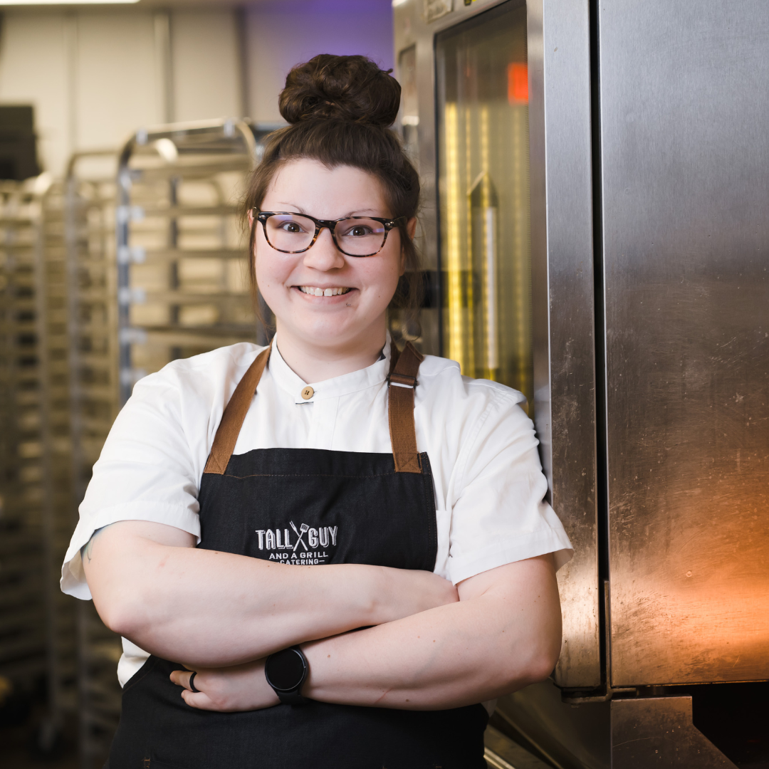 A smiling woman with glasses, wearing a white chef's coat and black apron, standing with arms crossed in a commercial kitchen.