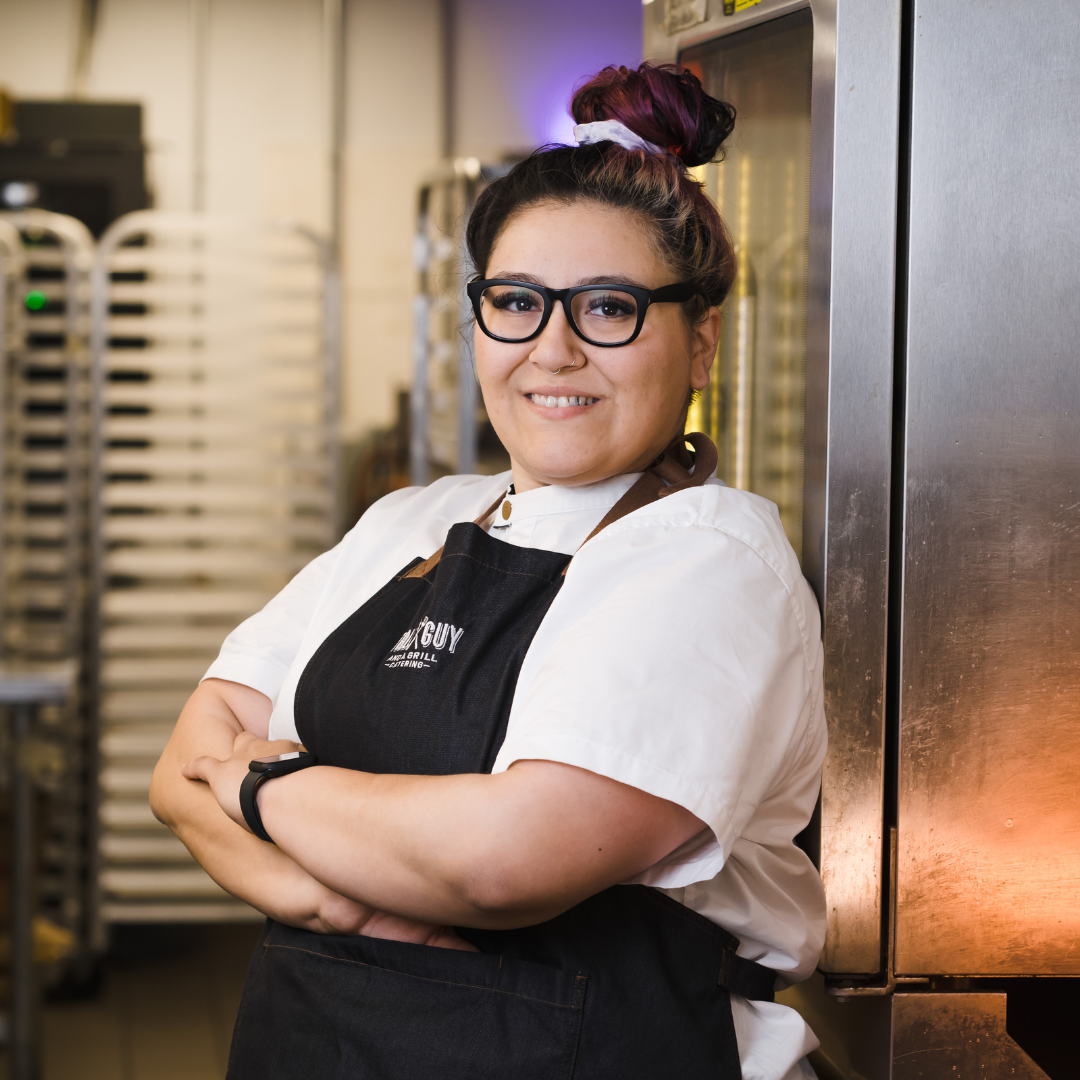 A woman with glasses and colorful hair tied up in a bun, smiling and standing with arms crossed in a commercial kitchen, wearing a white chef's coat and black apron.