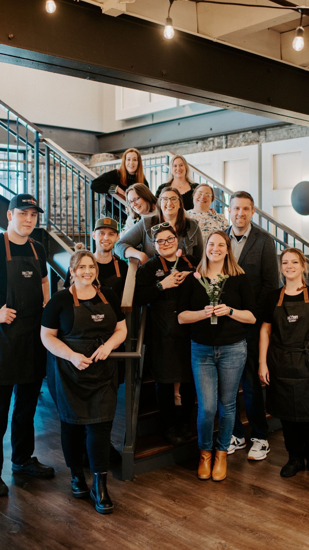 Group of people, some wearing Tallyguy aprons, posing for a photo inside a restaurant or cafe. Two women in the front are holding flowers, one with a small bouquet and another with a single bloom. The group is smiling, standing on and around a staircase with black railings.