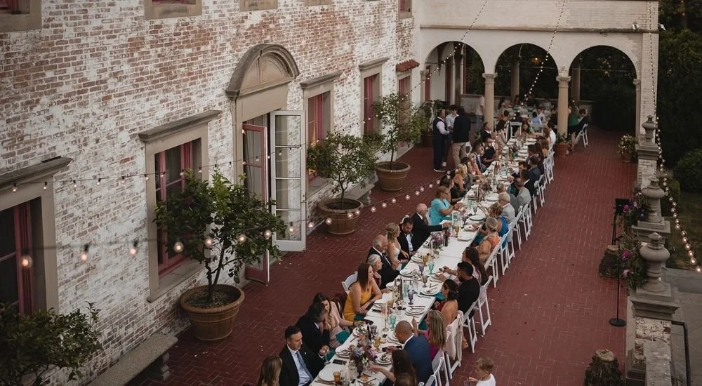 Long banquet table set outdoors on a brick terrace with guests dining, string lights, potted trees, and a brick building wall with open windows in the background.