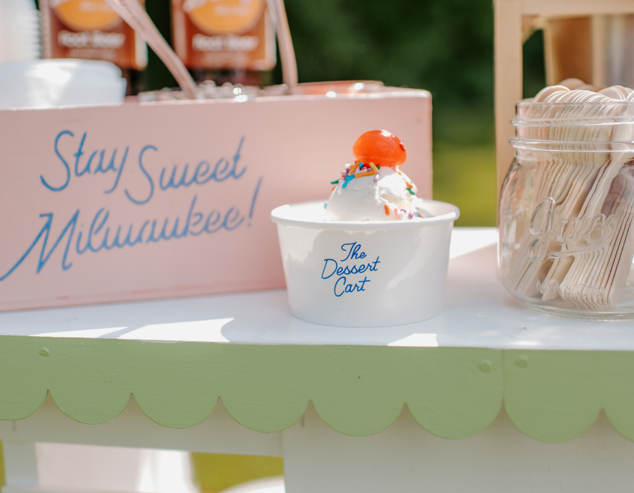 A bowl of vanilla ice cream with colorful sprinkles and a cherry on top, placed on a white table with a sign that says 'Stay Sweet Milwaukee!' in the background and a jar of wooden utensils nearby.