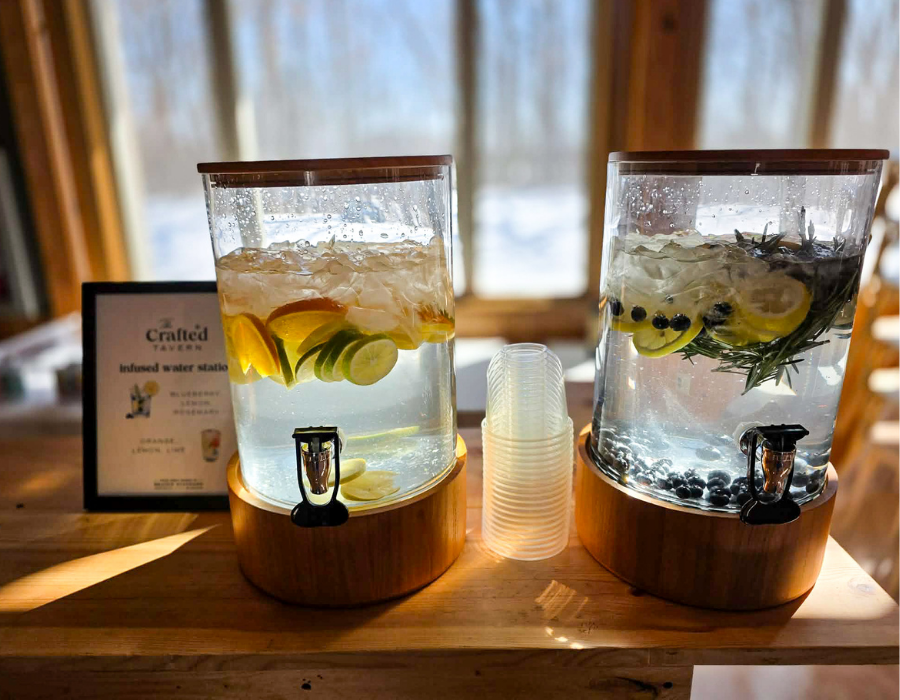 Two glass dispensers with flavored water, one with lemon and lime slices, the other with lemon slices, blueberries, and rosemary sprigs, on a wooden counter with cups in the middle.