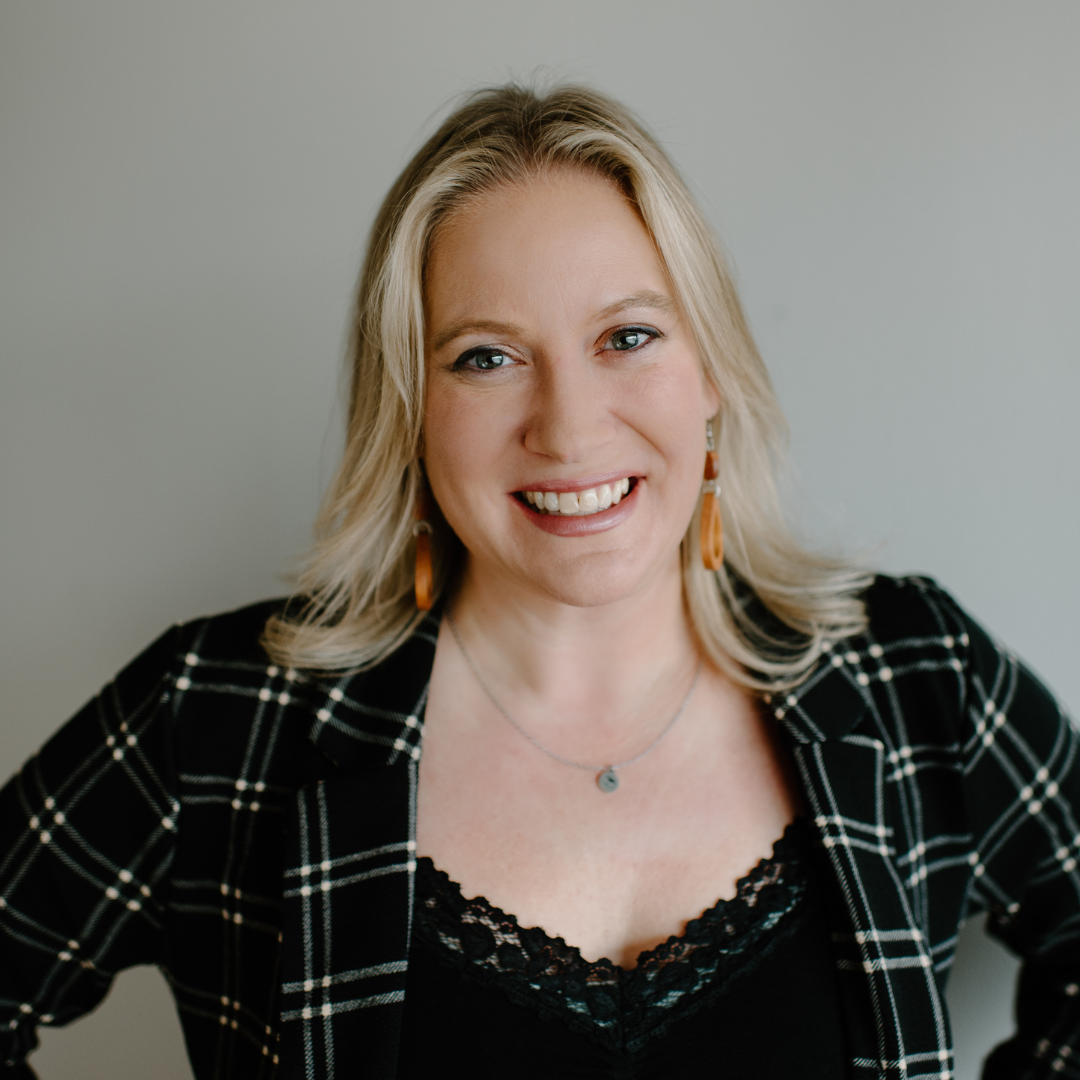 A woman with blonde hair smiling at the camera, wearing a black lace top and a black and white checkered blazer, with orange earrings and a small pendant necklace, standing against a plain light gray background.