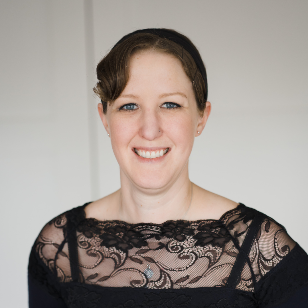A woman with short brown hair, blue eyes, and a nose piercing, smiling, wearing a black lace top with floral patterns.