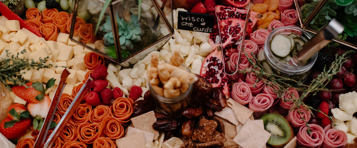 An arrangement of cheeses, fruits, and snacks at a charcuterie board, including strawberries, raspberries, kiwi, and various types of cheese, with some labeled items and garnishes.
