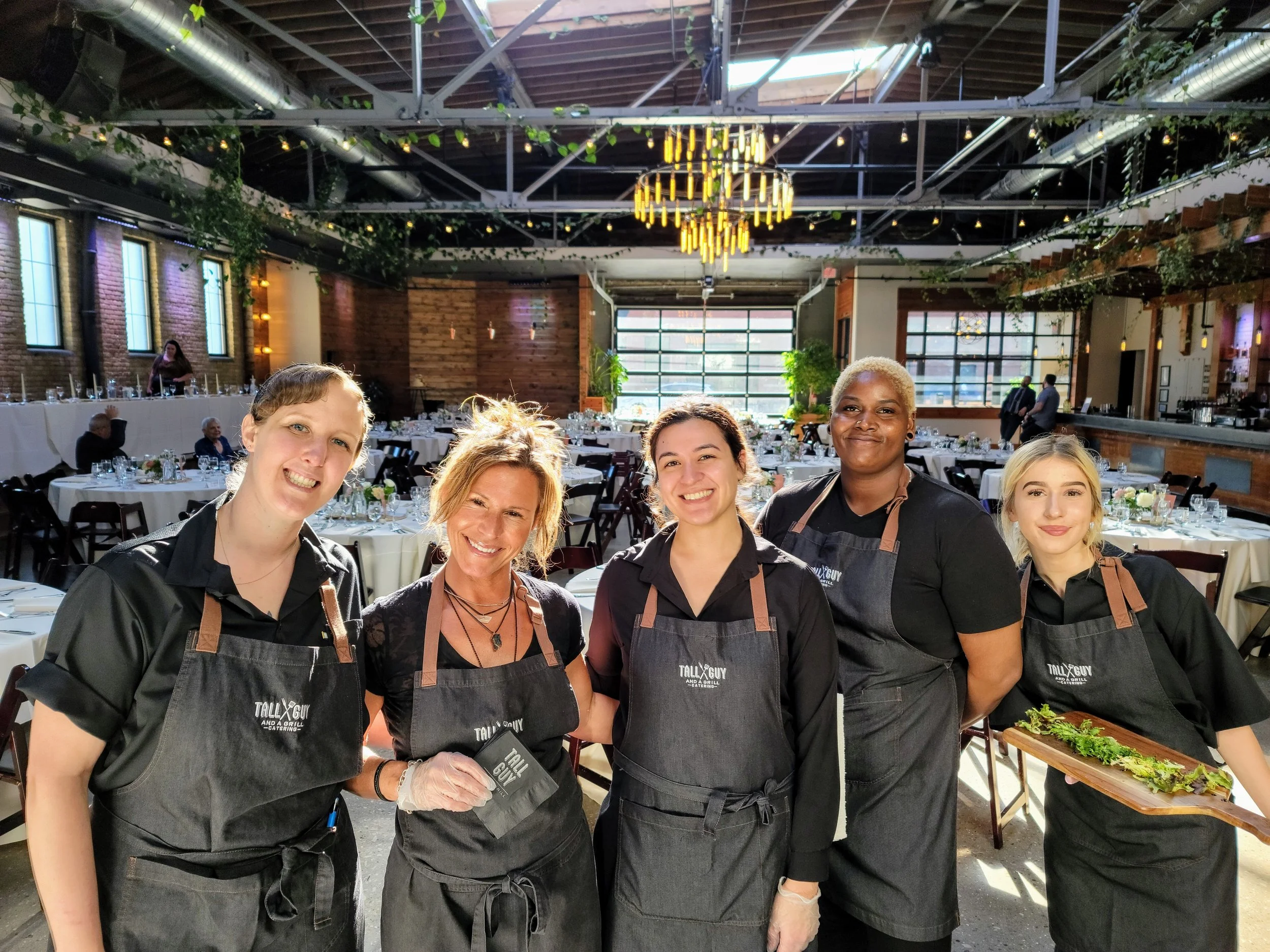 Five women in black shirts and gray aprons posing inside a decorated banquet hall with tables set for an event.