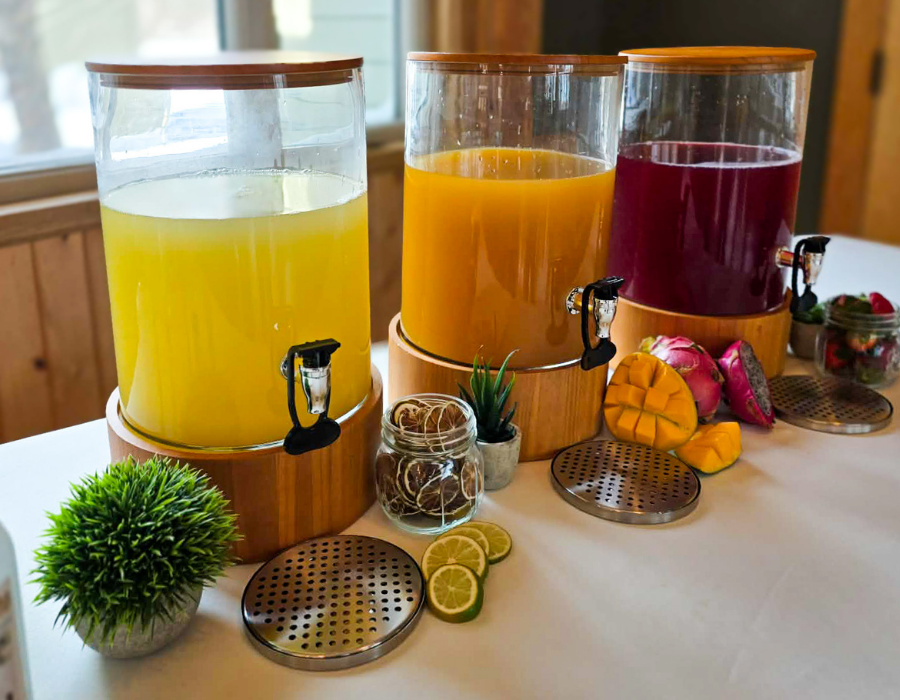 Three large beverage dispensers filled with yellow, orange, and purple drinks on a table, with lemon slices, mango, sweet potatoes, strawberries, and decorative plants nearby.