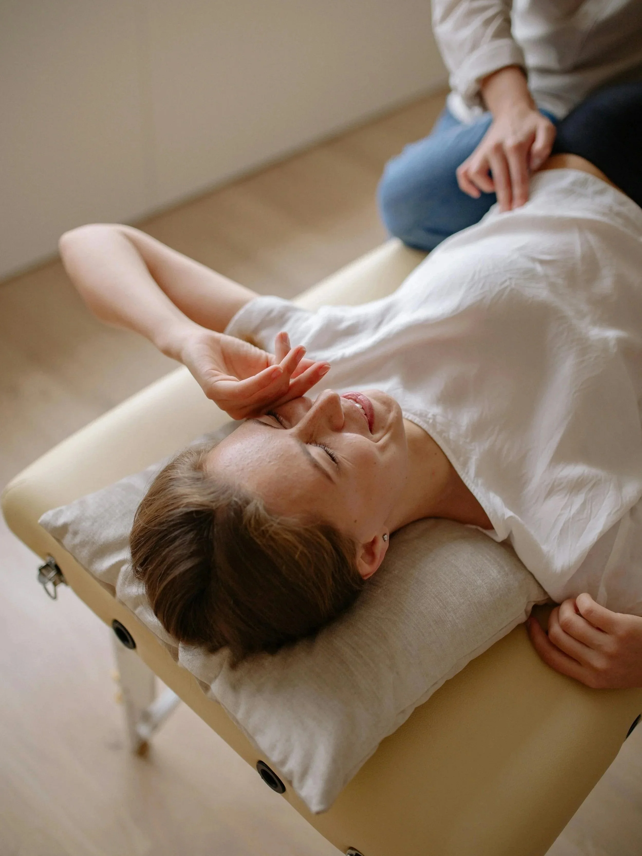 A woman lying on a massage table with her eyes closed, receiving a facial massage from a practitioner.
