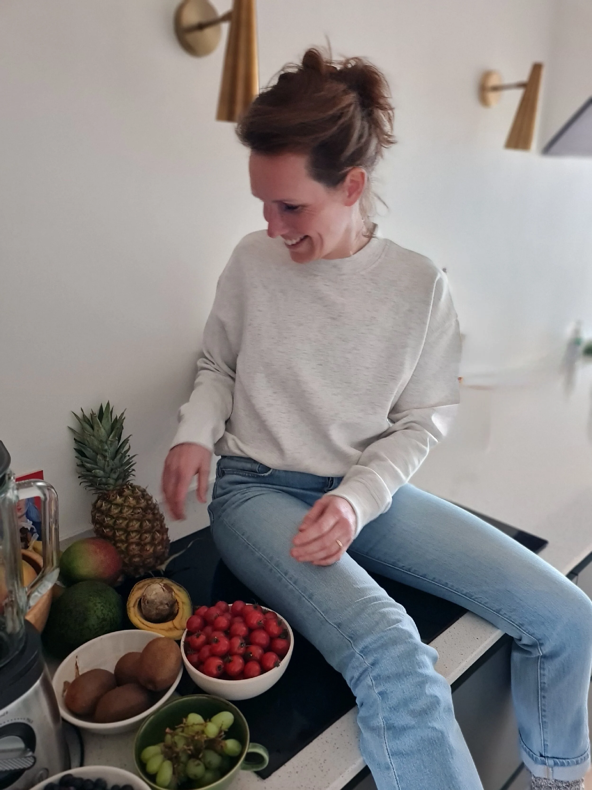 Vrouw zittend op het aanrecht in de keuken, omringd door verschillende soorten fruit zoals ananas, kiwi's, kersen en druiven, lachend en genietend.