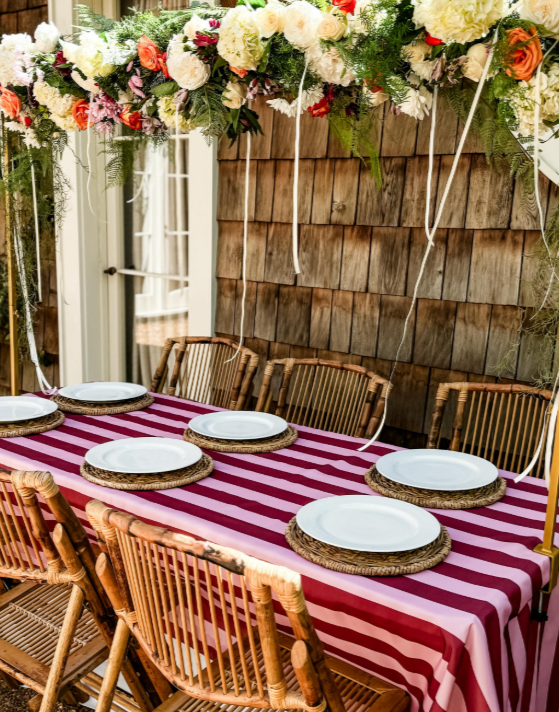 Outdoor dinner table set with a striped cloth, woven chargers and a floral garland overhead