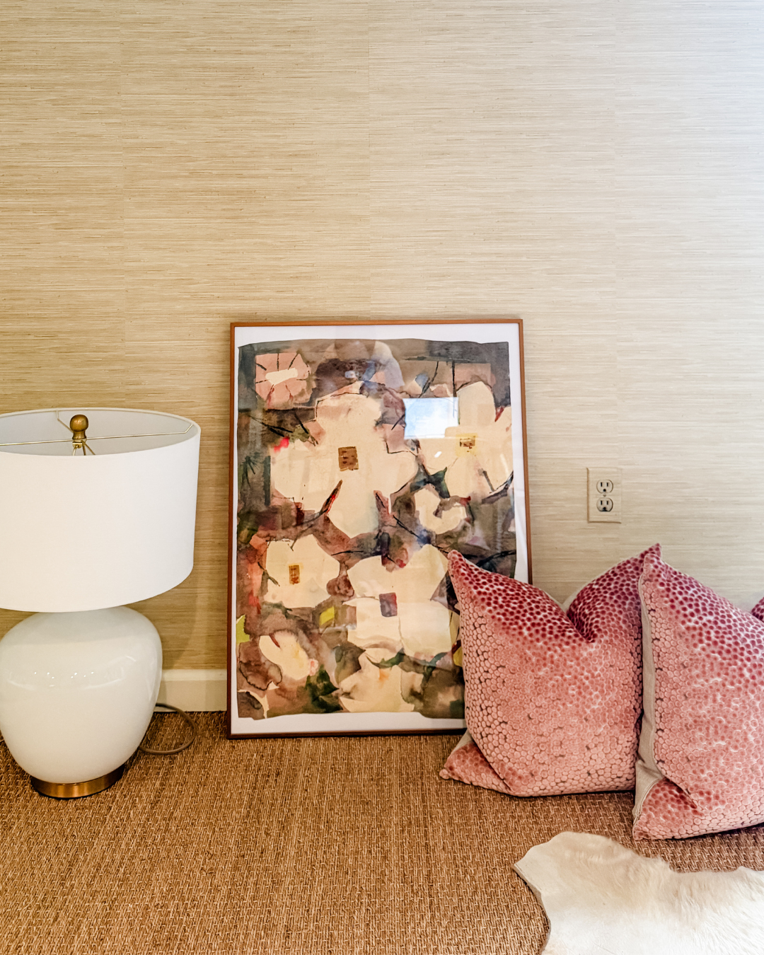 Abstract framed art leaning against a grasscloth wall with a white lamp and pink pillows