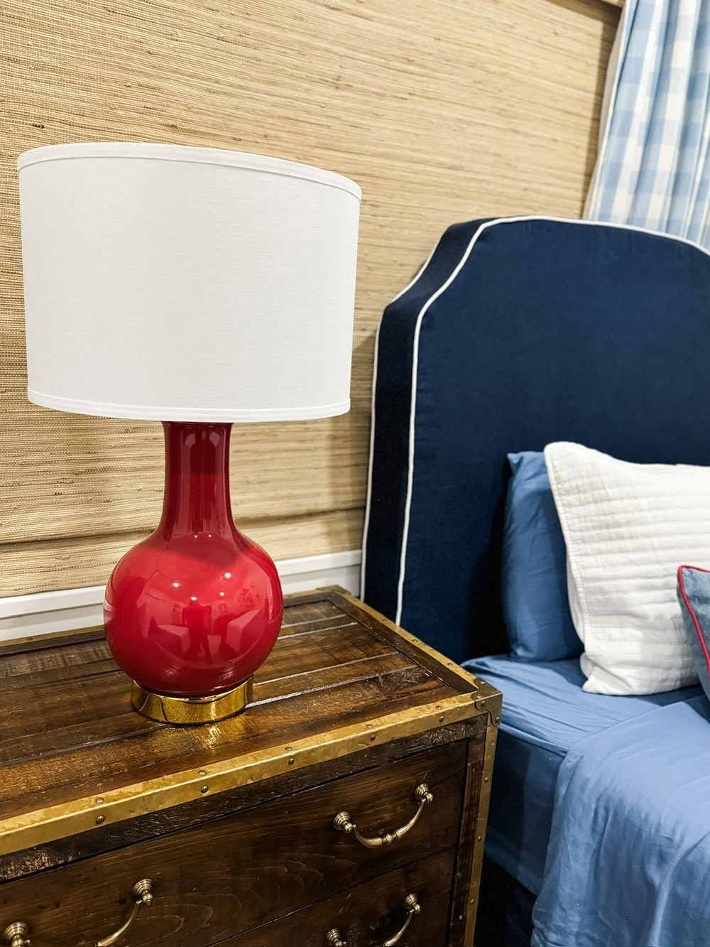 Red ceramic lamp on a wooden dresser beside a navy upholstered headboard and blue dorm bedding