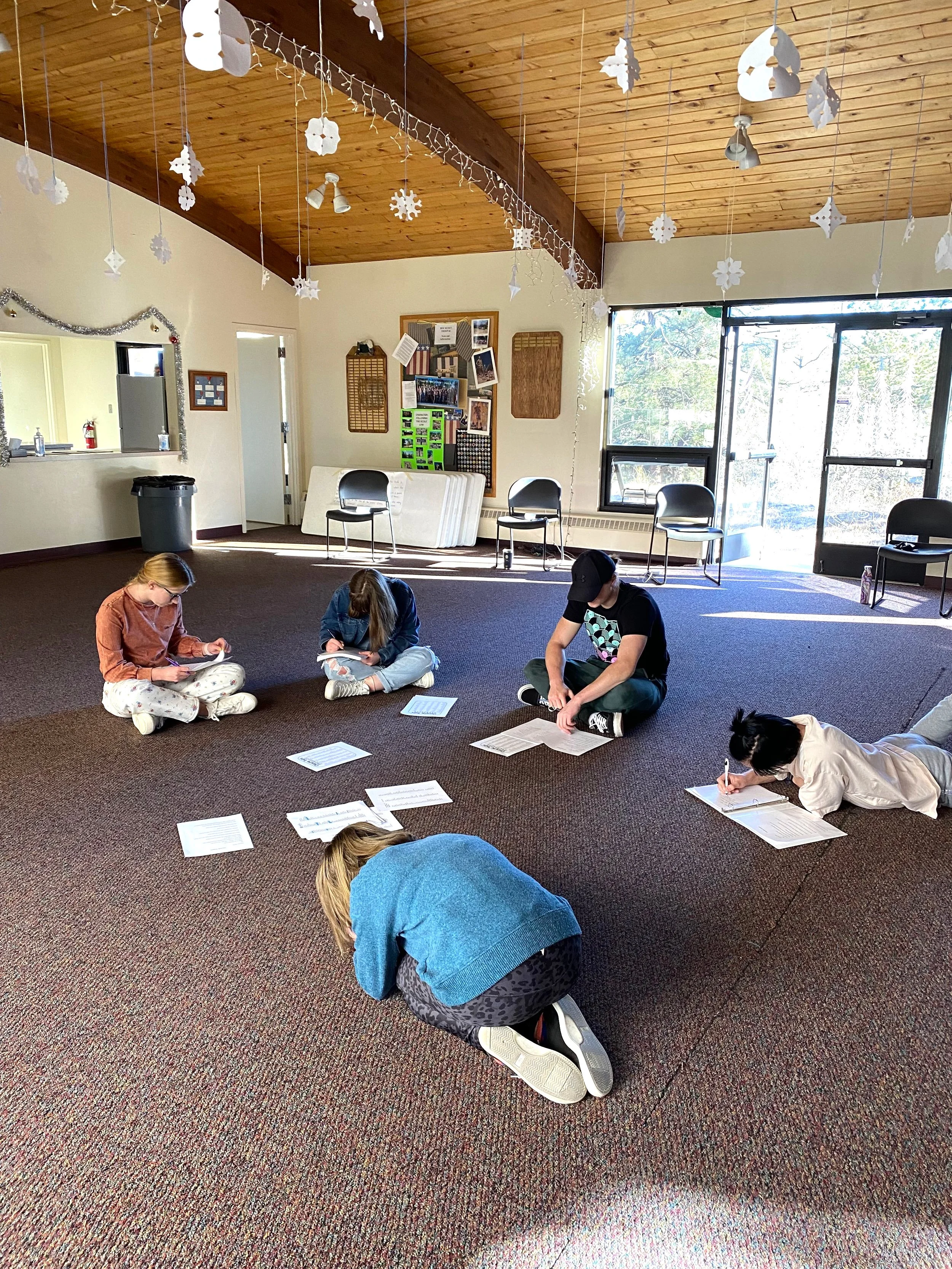 Group of people sitting on the floor in a room with wooden ceiling and paper snowflakes hanging. They are writing on papers spread on the floor.