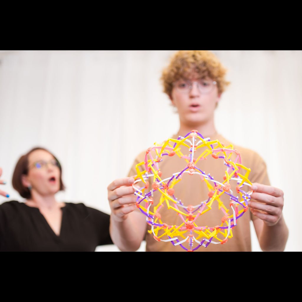 Two people in a room, one holding a colorful geometric structure, looking surprised.