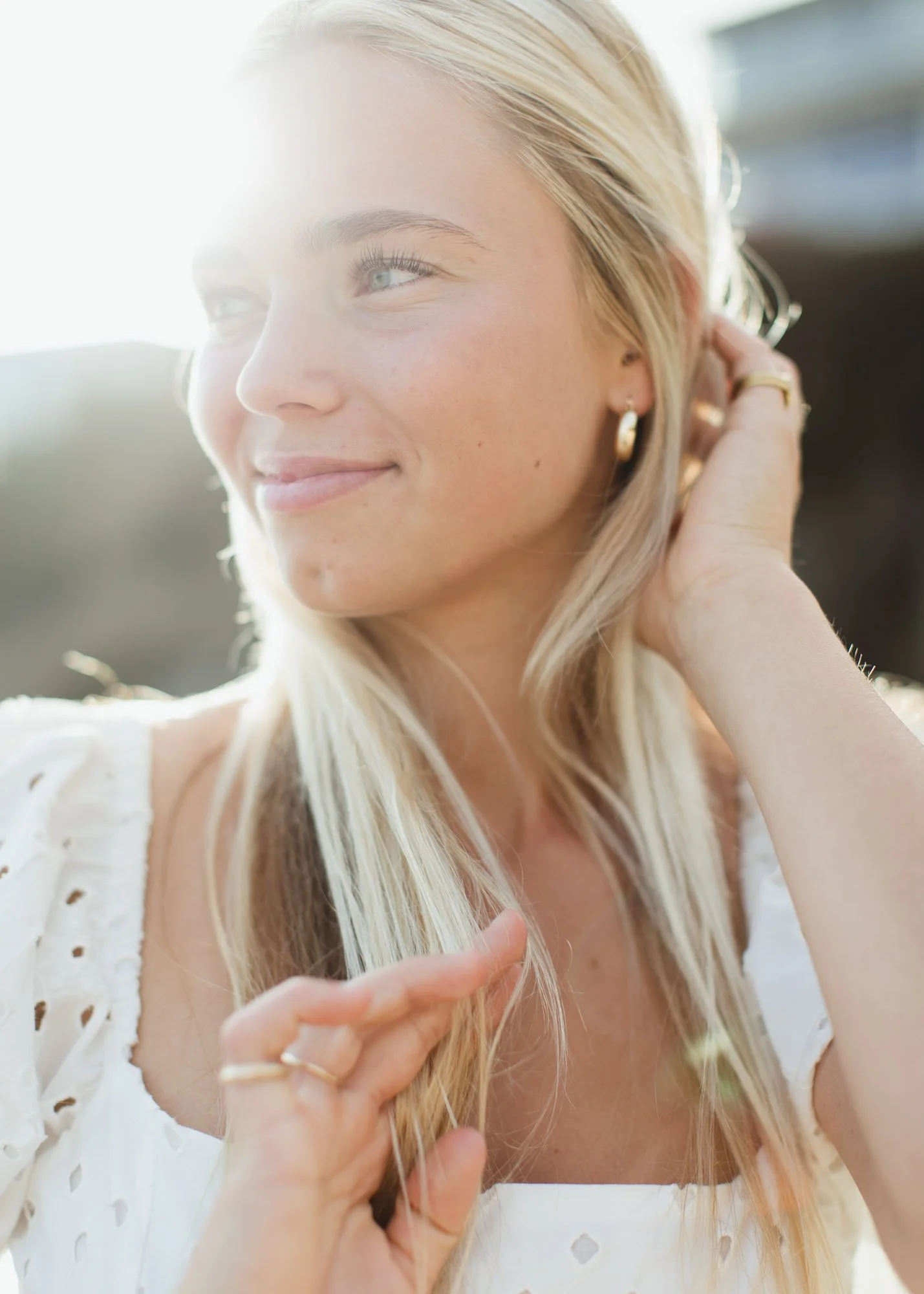 A photo of a high school senior girl with long blonde hair, smiling softly and looking off to the side, with sunlight illuminating her face, wearing a white eyelet top and gold hoop earrings.