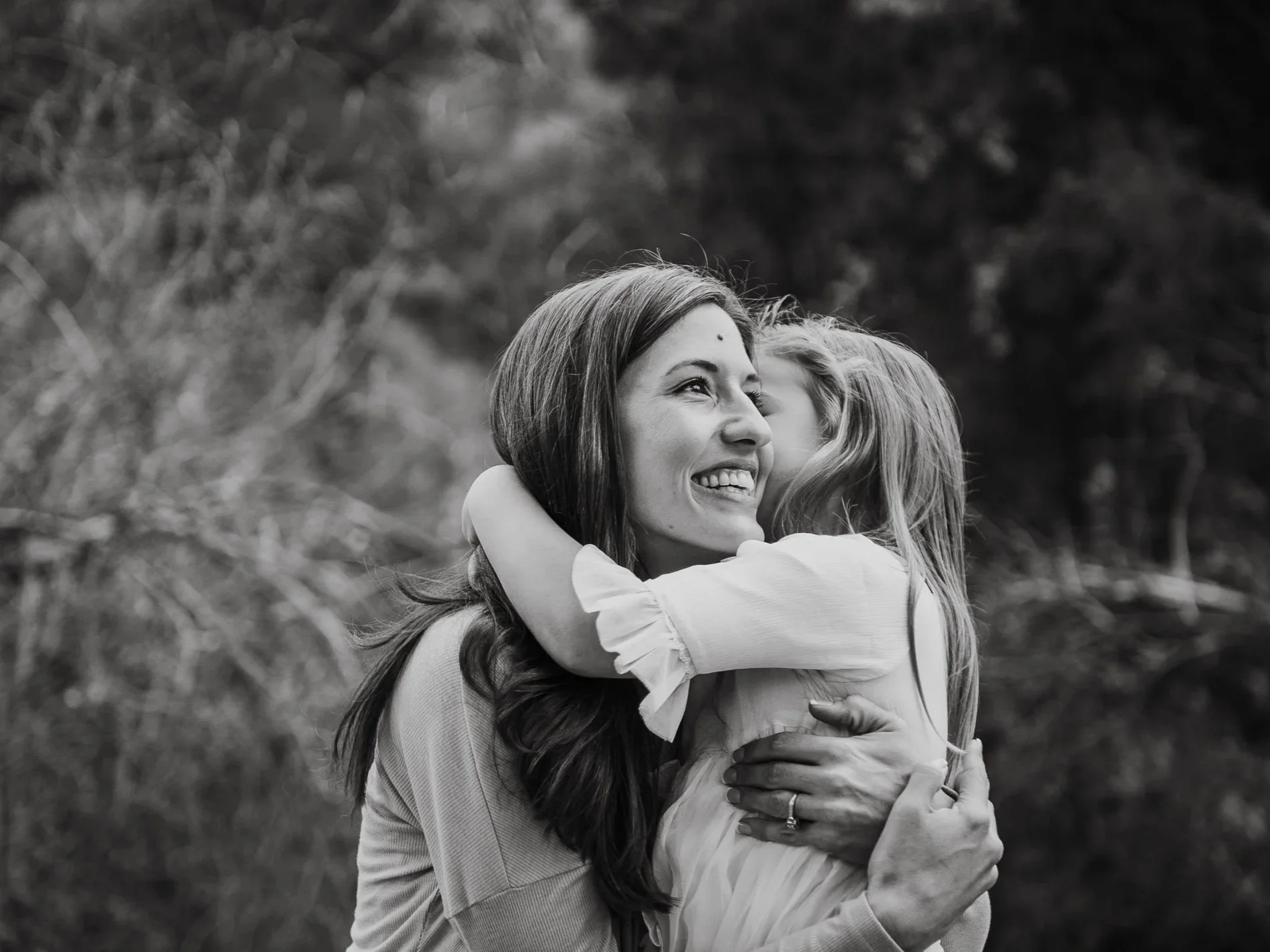 Mother with long hair smiling and hugging her daughter with long hair, possibly outdoors in a natural setting, in black and white.