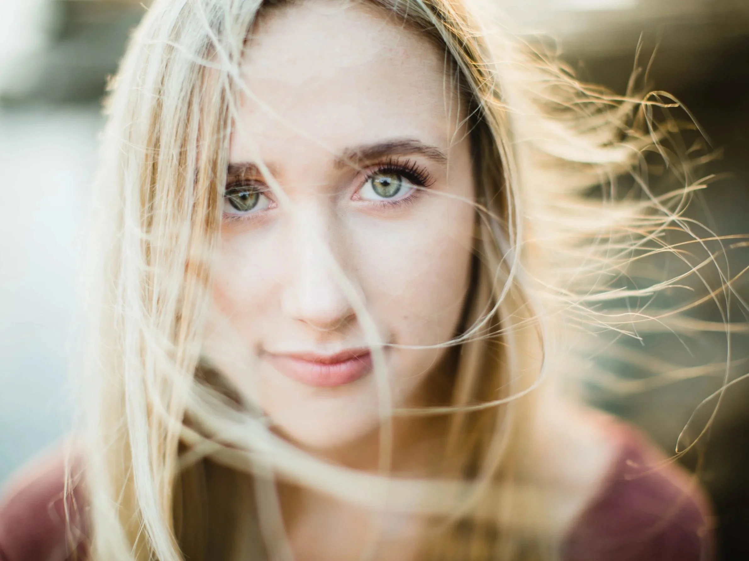 A close-up of a high school senior girl with green eyes and blonde hair, outdoors, with sunlight and windmoving her hair.