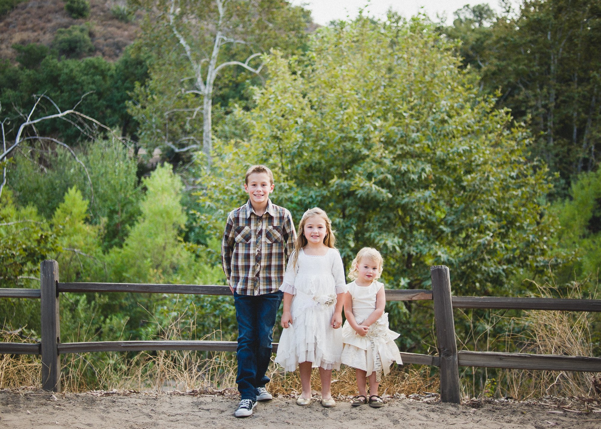 Orange County Child and Family Photography. Three children standing in front of a wooden fence with a forest background, wearing casual clothing.