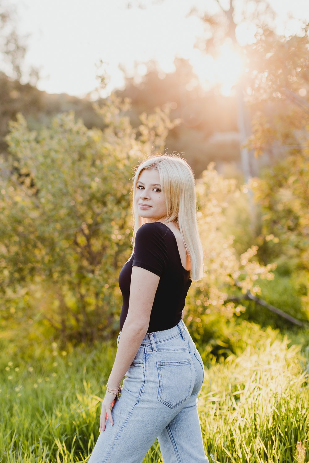 High school senior portrait girl with long blonde hair wearing a black top and jeans standing in a sunlit field with green foliage.