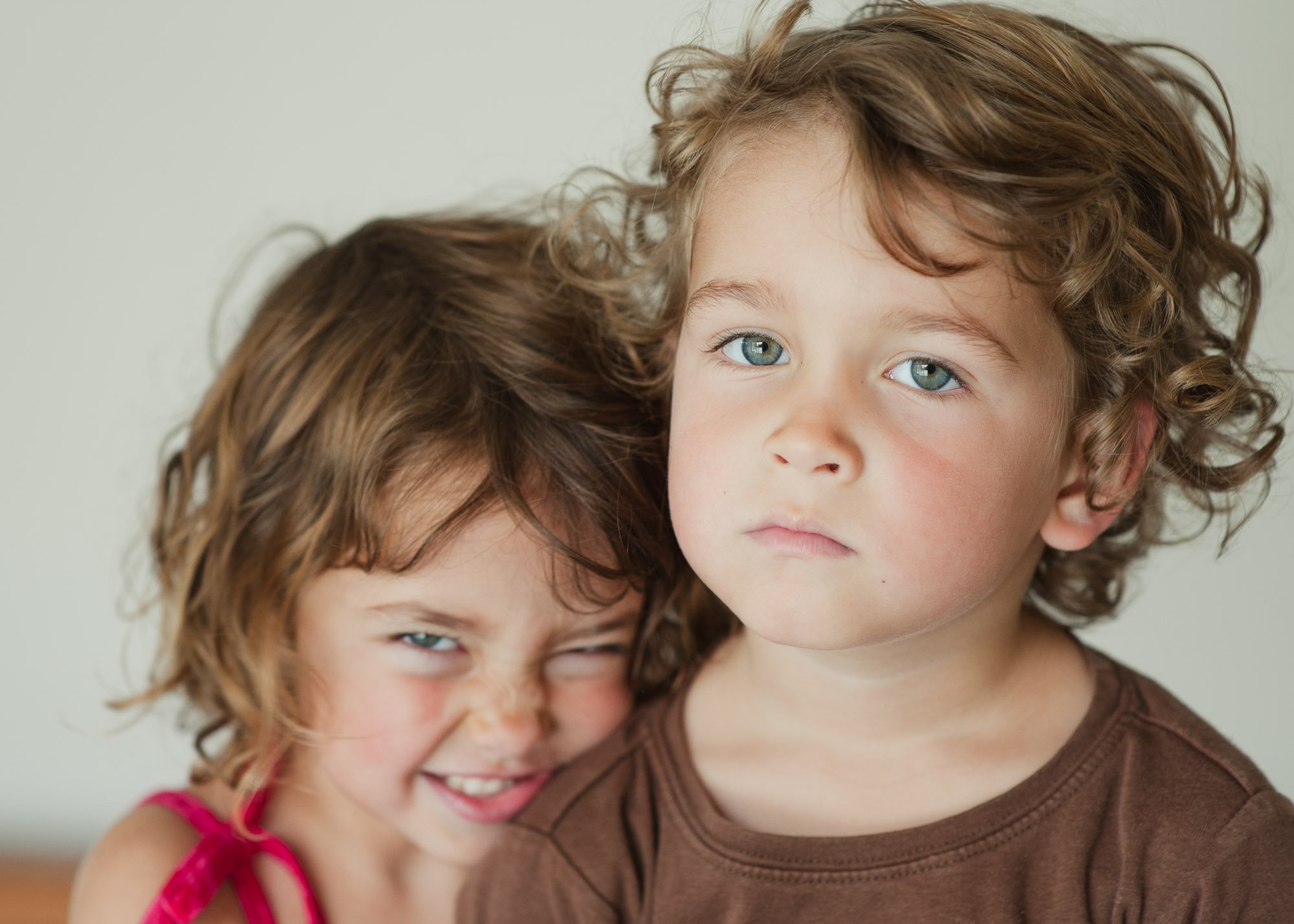 Orange County Child and Family Photography. Two young children with curly hair; one smiling playfully, the other looking serious, indoors against a neutral background.