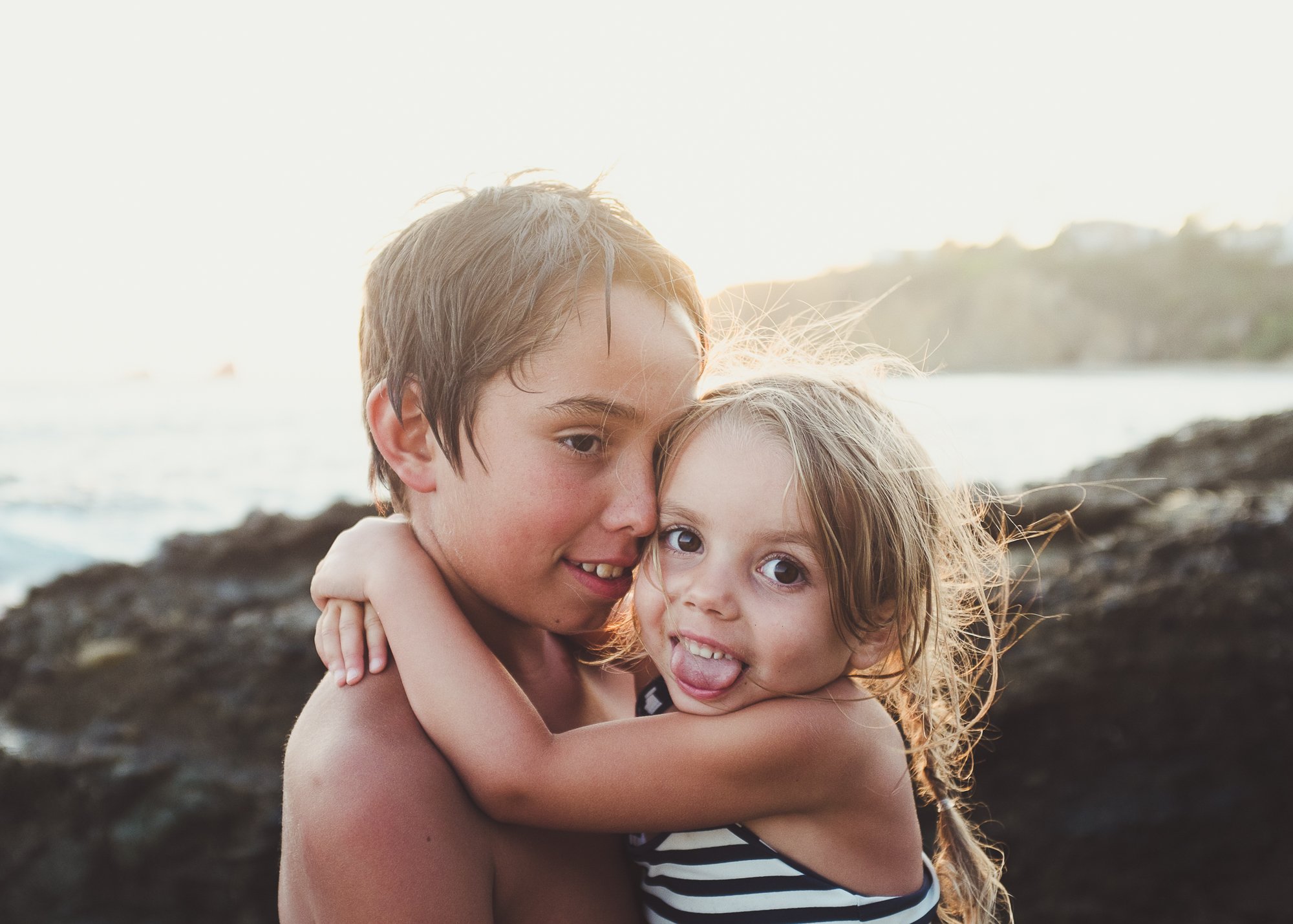 Orange County Child and Family Photography. Two children hugging at the beach, one smiling and the other sticking out their tongue, with sunlight in the background.