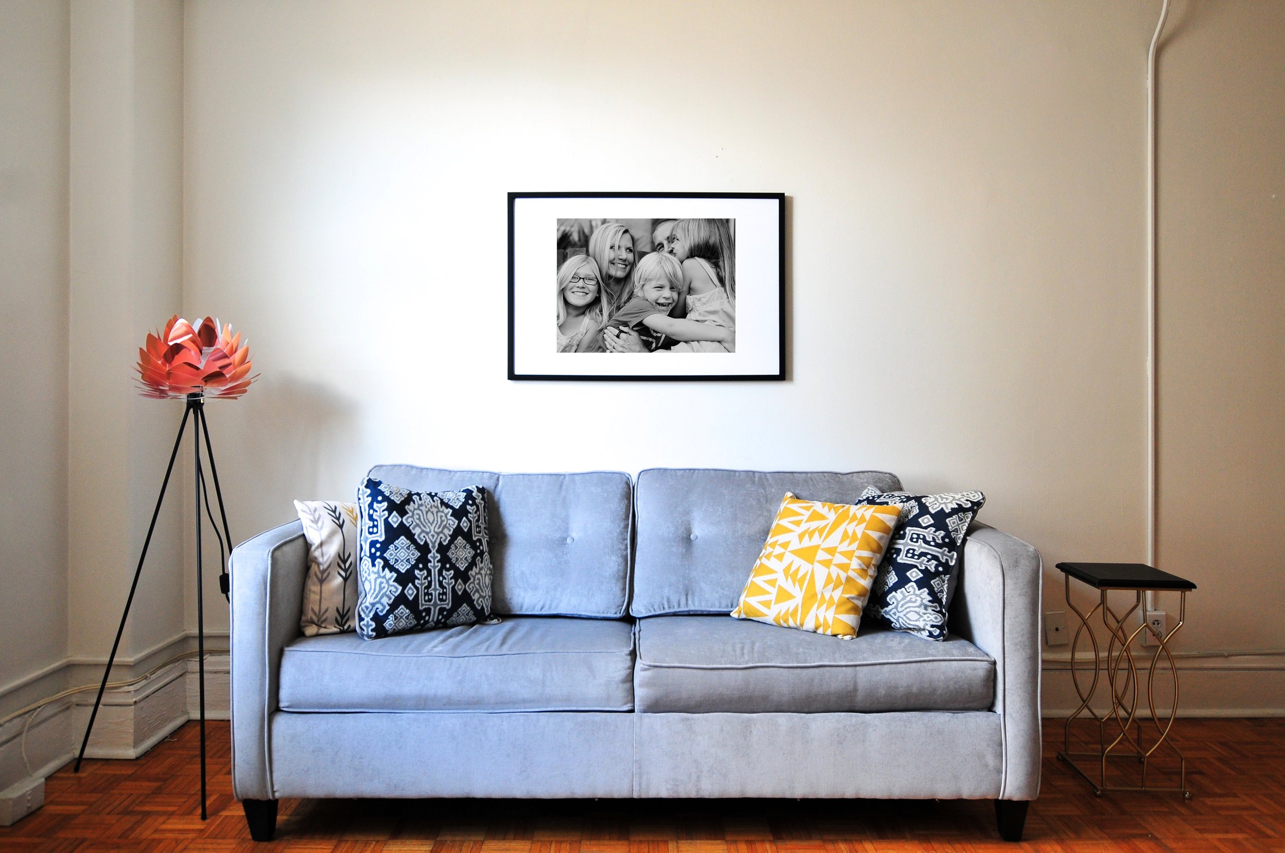 Modern living room interior with gray sofa, colorful cushions, framed black-and-white photo on wall, tripod floor lamp with red lampshade, and small side table on wooden floor